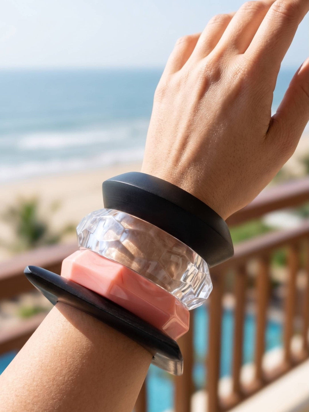 Four stacked acrylic bangles on a wrist held up against a sunny beach backdrop
