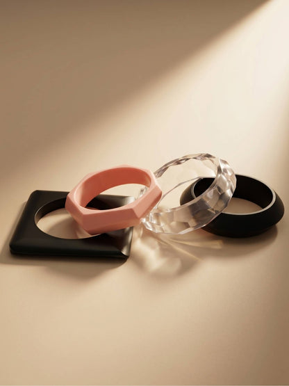 Four acrylic bangles laid flat — black square, pink geometric, clear faceted, black oval — on a warm-toned surface