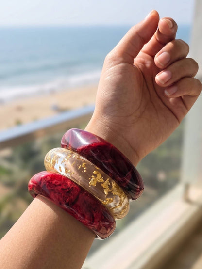 Three stacked resin bangles on a raised wrist against a beachside backdrop, two crimson marble and one clear with gold foil