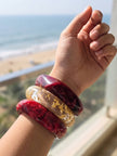 Three stacked resin bangles on a raised wrist against a beachside backdrop, two crimson marble and one clear with gold foil