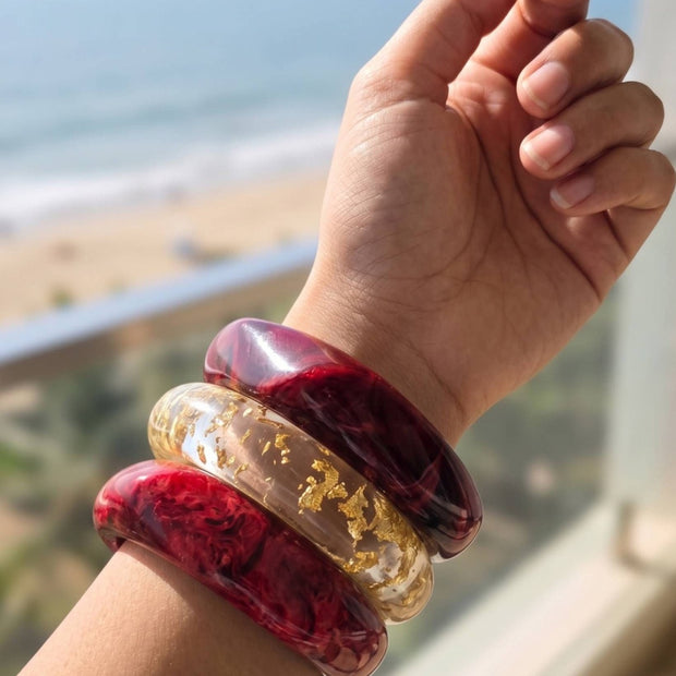 Three stacked resin bangles on a raised wrist against a beachside backdrop, two crimson marble and one clear with gold foil