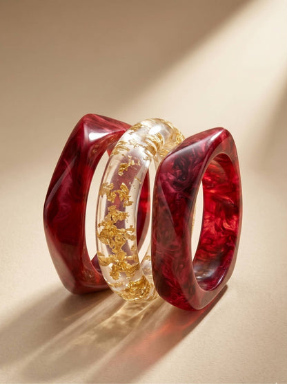 Set of three resin bangles displayed on a neutral surface, showing crimson marble swirl and transparent gold foil designs