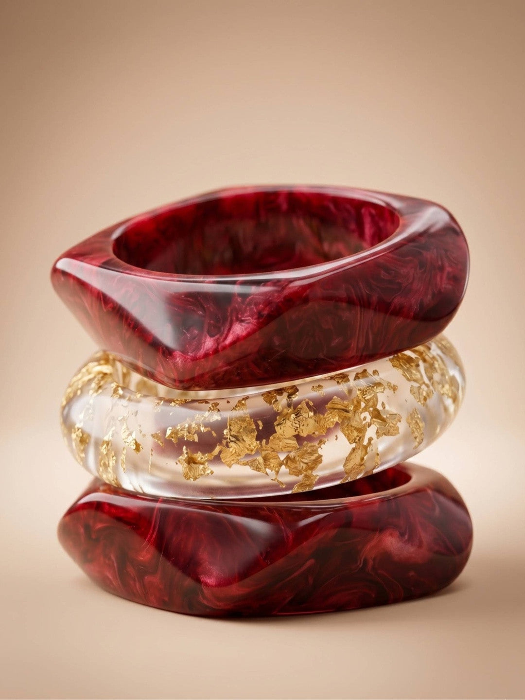 Three resin bangles stacked vertically on a beige surface — one angular crimson, one round gold foil, one round crimson marble