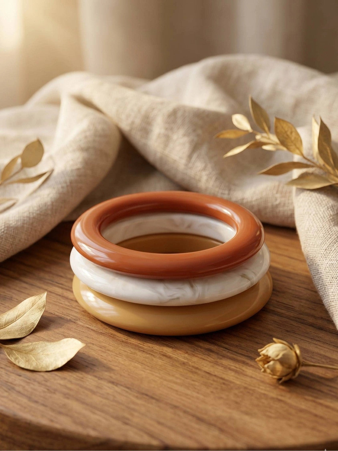 Three stacked resin bangles in terracotta, ivory and mustard on a wooden surface with dried golden leaves and linen fabric