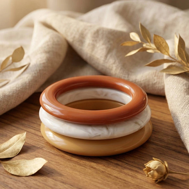Three stacked resin bangles in terracotta, ivory and mustard on a wooden surface with dried golden leaves and linen fabric