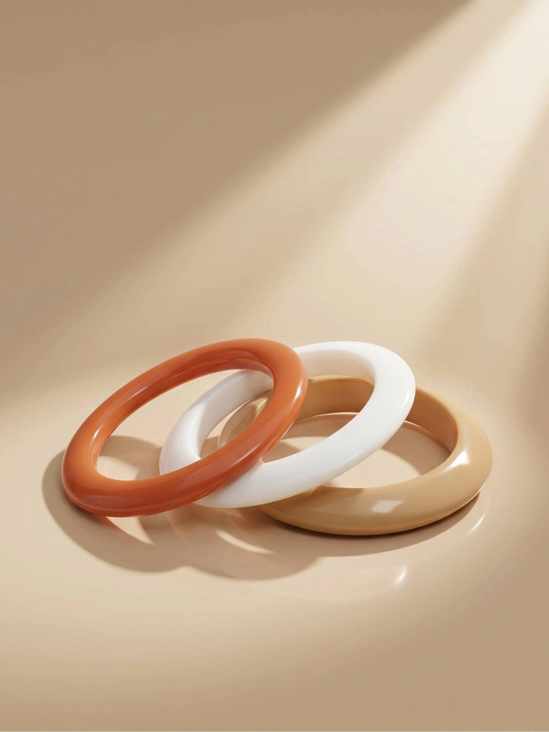 Three glossy resin bangles in terracotta, white and mustard leaning against each other on a beige surface in soft directional light