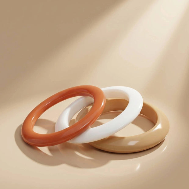 Three glossy resin bangles in terracotta, white and mustard leaning against each other on a beige surface in soft directional light