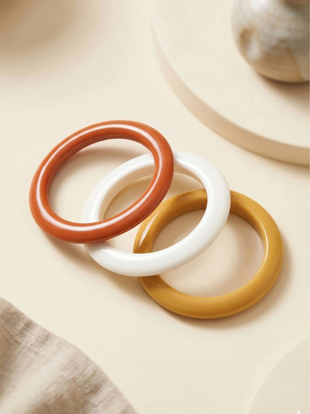 Three resin bangles in terracotta, ivory white and mustard laid flat and fanned out on a cream surface beside a linen cloth