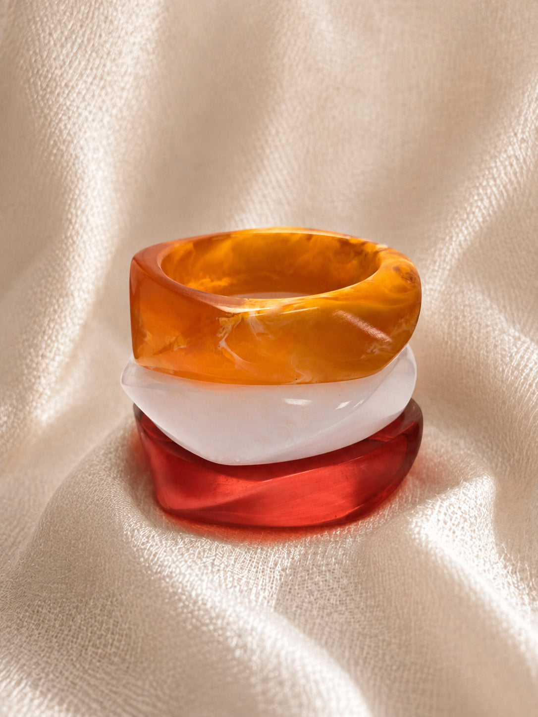 Amber, white and red chunky resin bangles stacked on top of each other on a cream fabric background