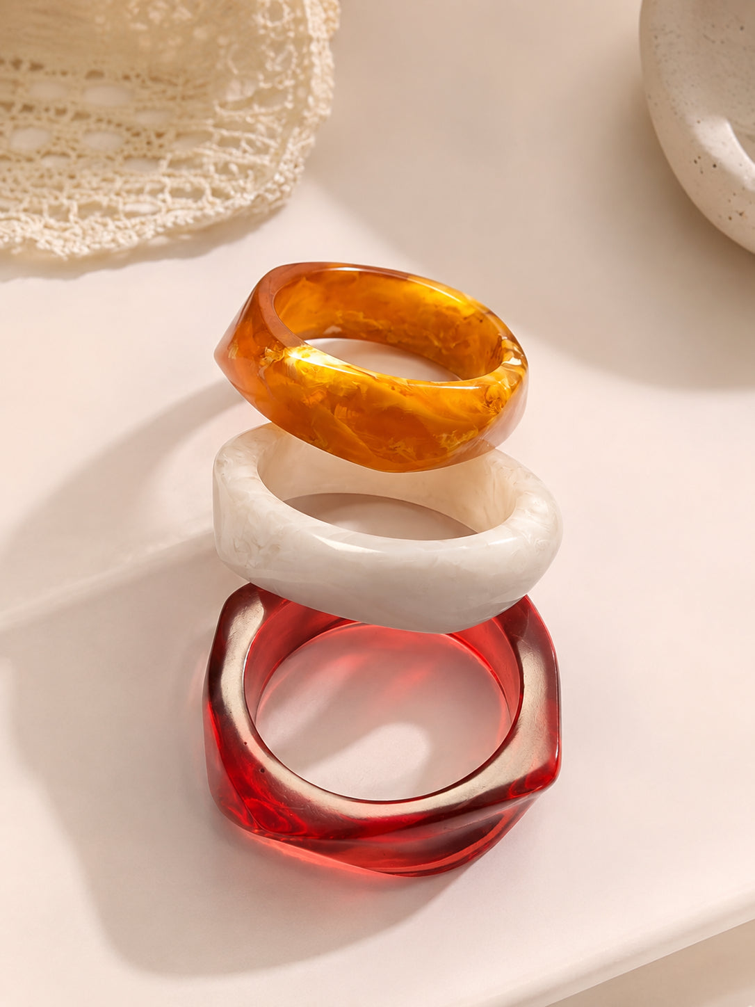 Amber, white and red resin bangles stacked in a tower on a white surface near lace fabric