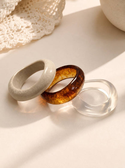 Three resin bangles in marble white, amber tortoiseshell and clear acrylic laid flat on a cream surface near lace