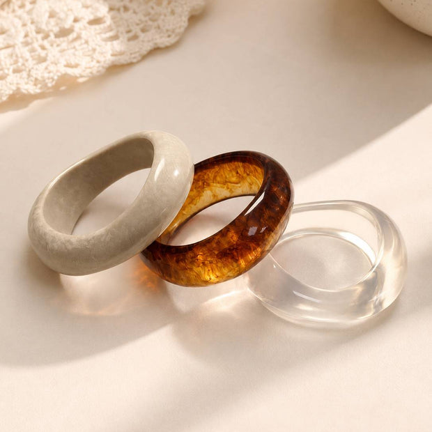 Three resin bangles in marble white, amber tortoiseshell and clear acrylic laid flat on a cream surface near lace