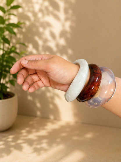 Model wearing all three resin bangles together on wrist — marble white, amber tortoiseshell and clear acrylic