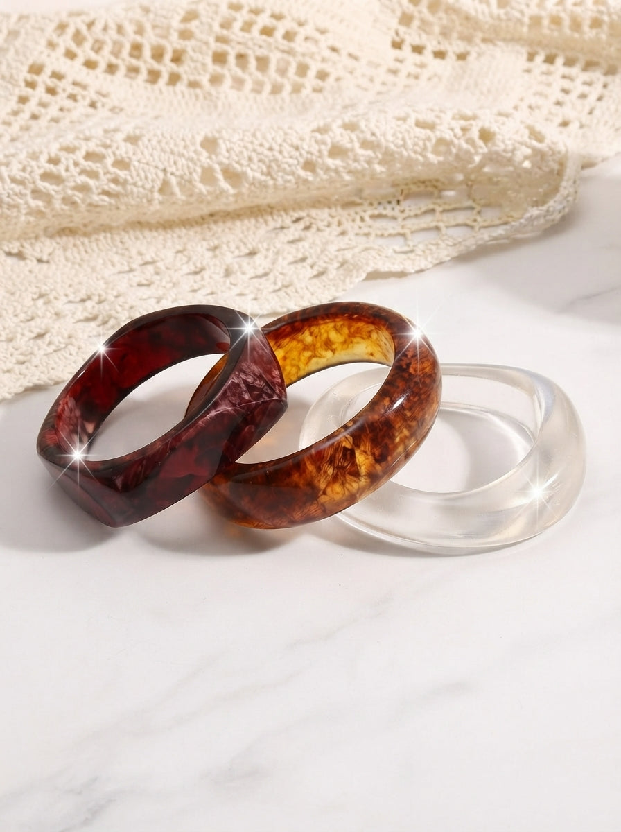 Three resin marble bangles in burgundy, tortoiseshell amber and clear crystal laid on white marble beside lace fabric