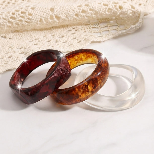 Three resin marble bangles in burgundy, tortoiseshell amber and clear crystal laid on white marble beside lace fabric
