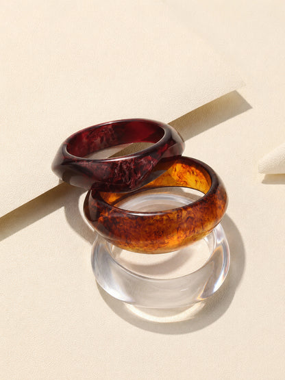 Three stacked resin bangles in burgundy, amber tortoiseshell and transparent clear on a cream textured surface