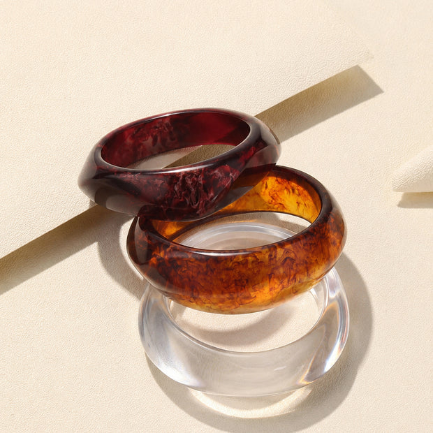 Three stacked resin bangles in burgundy, amber tortoiseshell and transparent clear on a cream textured surface