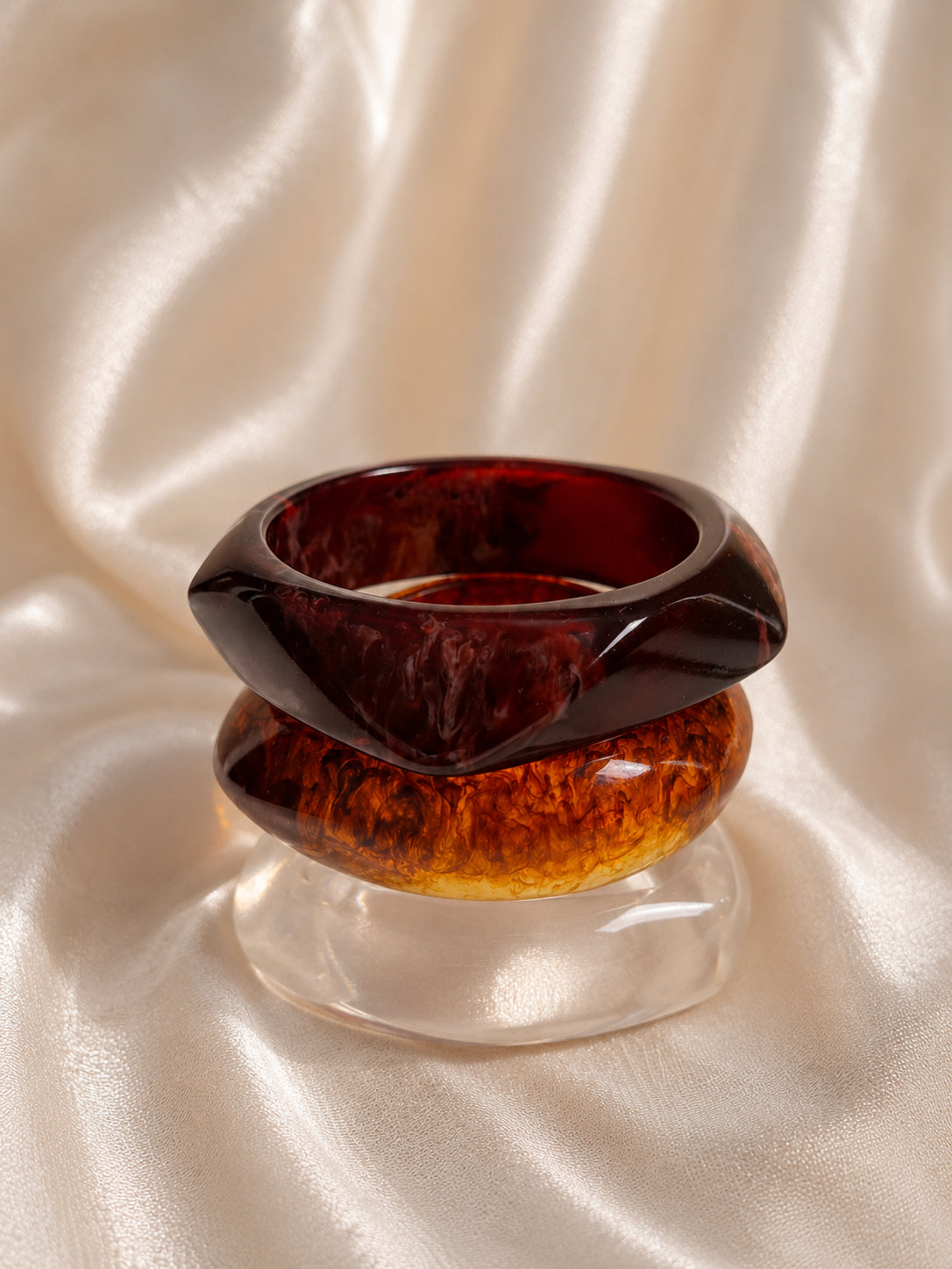 Close-up of burgundy and amber tortoiseshell resin bangles stacked on a clear bangle against draped satin fabric