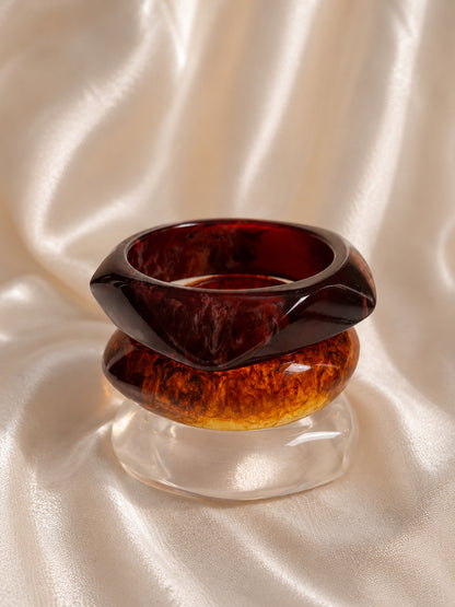 Close-up of burgundy and amber tortoiseshell resin bangles stacked on a clear bangle against draped satin fabric