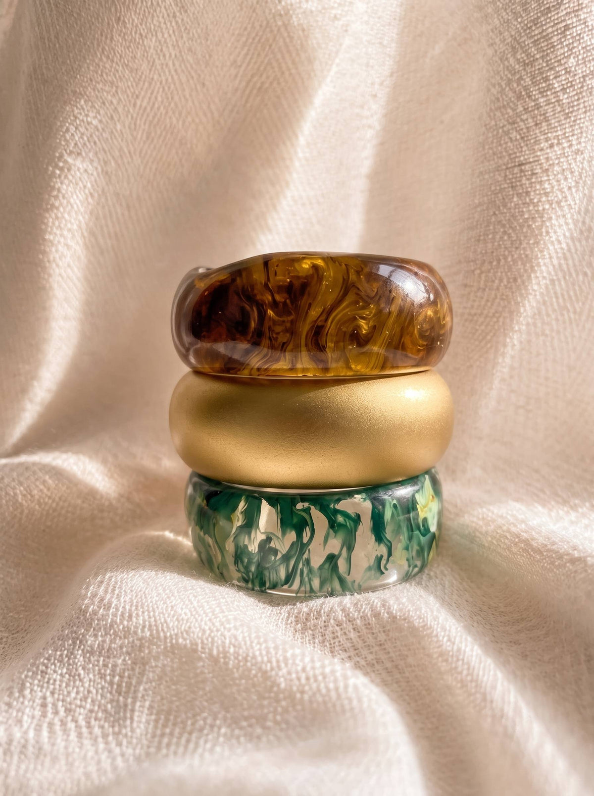Three resin bangles stacked vertically on cream fabric – amber marble on top, matte gold in middle, green marble at base