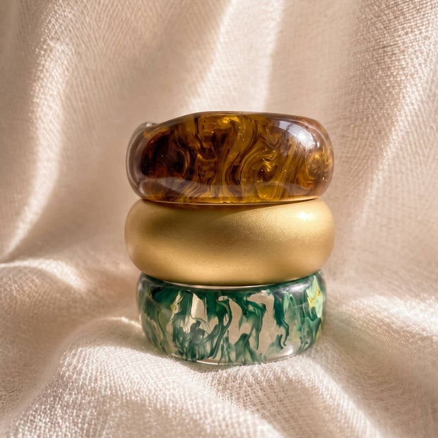 Three resin bangles stacked vertically on cream fabric – amber marble on top, matte gold in middle, green marble at base