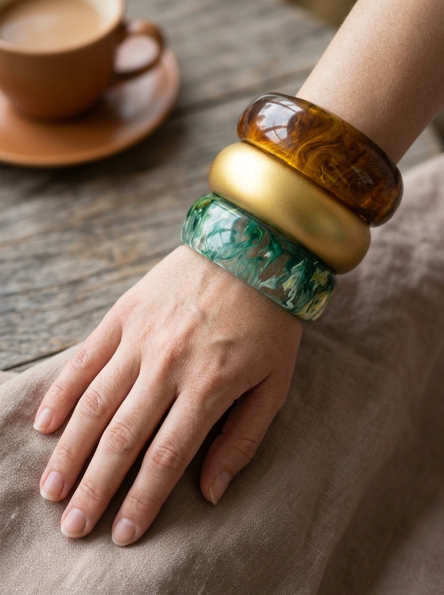 All three resin marble bangles worn stacked on a woman's wrist resting on a linen cloth with a coffee cup in background