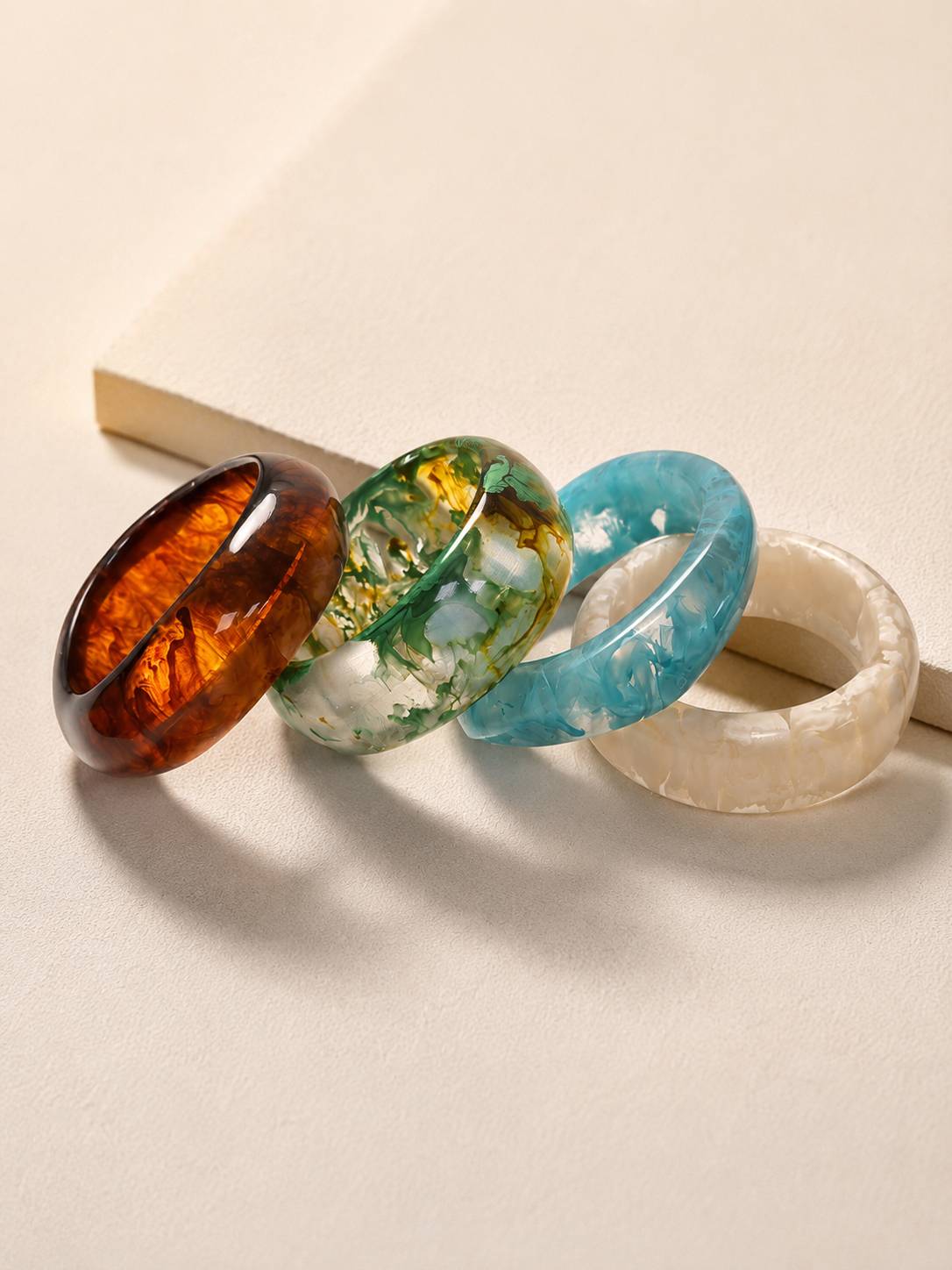 Four resin marble bangles laid side by side on a neutral surface displaying tortoiseshell, botanical green, ocean blue and ivory finishes