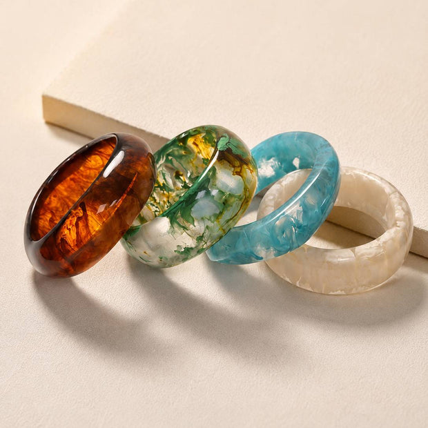 Four resin marble bangles laid side by side on a neutral surface displaying tortoiseshell, botanical green, ocean blue and ivory finishes