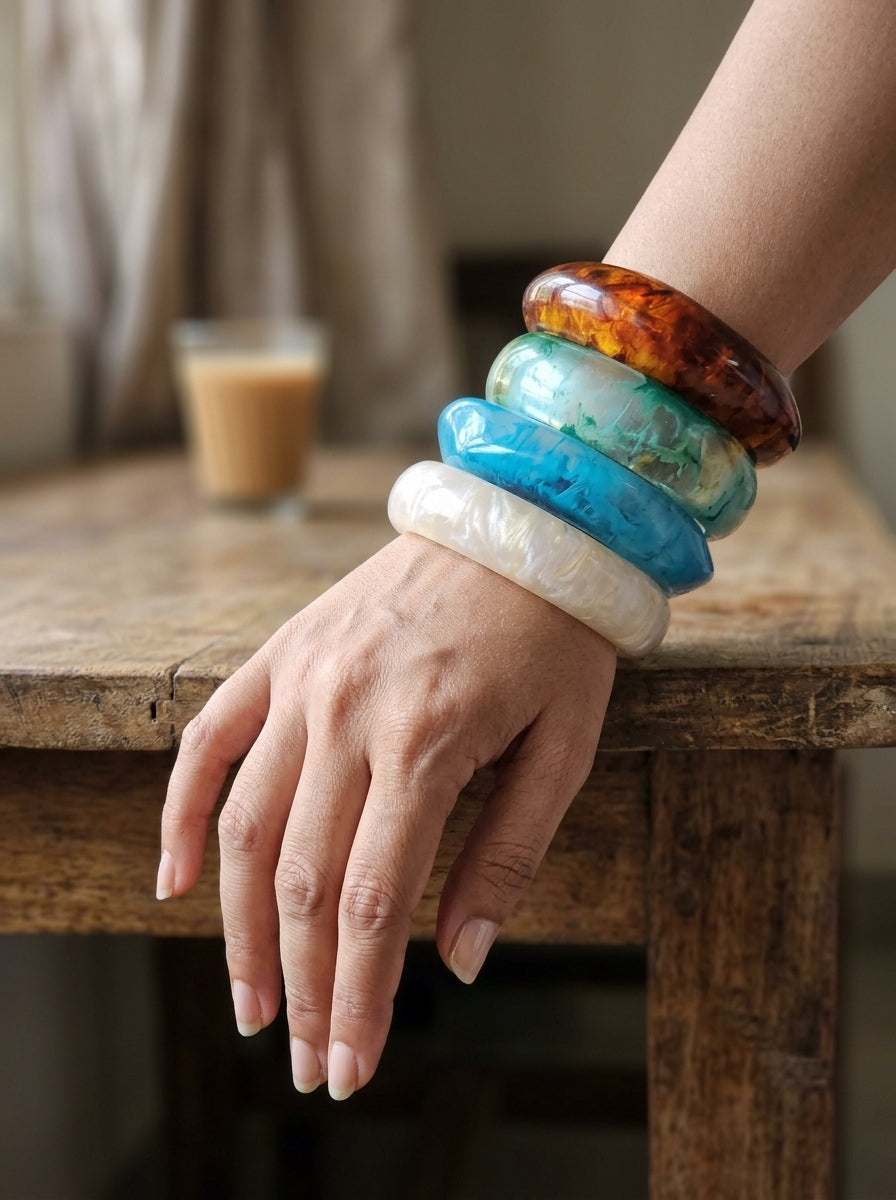 Model wearing all four stacked resin marble bangles on wrist resting on a wooden table with a coffee glass in background