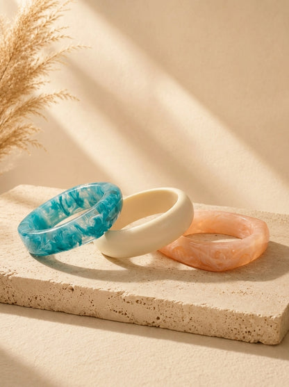 Three marble resin bangles in teal, ivory and peach laid flat on a textured stone surface with pampas grass in background