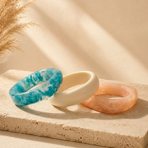 Three marble resin bangles in teal, ivory and peach laid flat on a textured stone surface with pampas grass in background