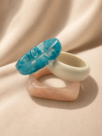 Teal marbled, ivory and blush peach resin bangles stacked together on a soft beige fabric background