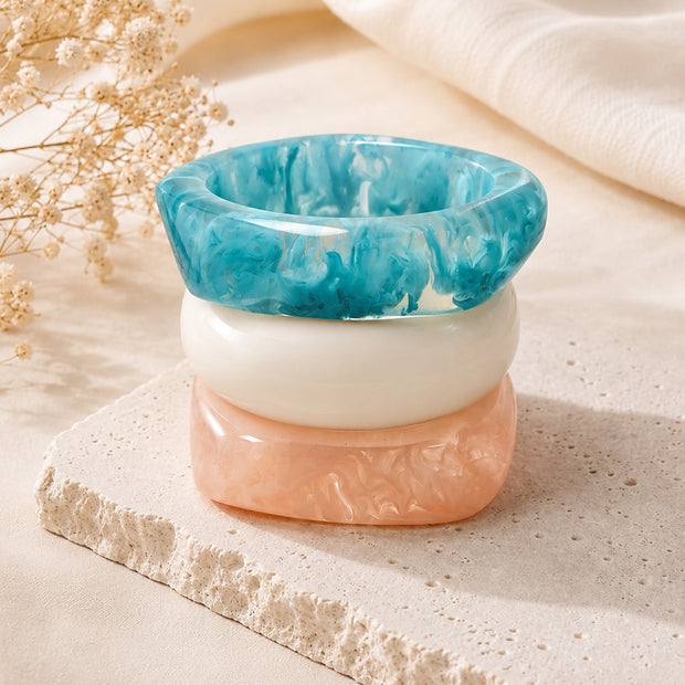Three marble resin bangles stacked upright on a stone riser showing teal, white and peach layers from above