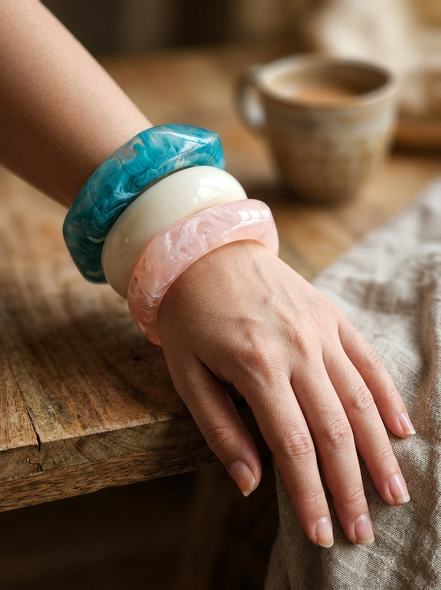 Woman wearing three stacked marble resin bangles in teal, ivory and peach on her wrist resting on a wooden surface