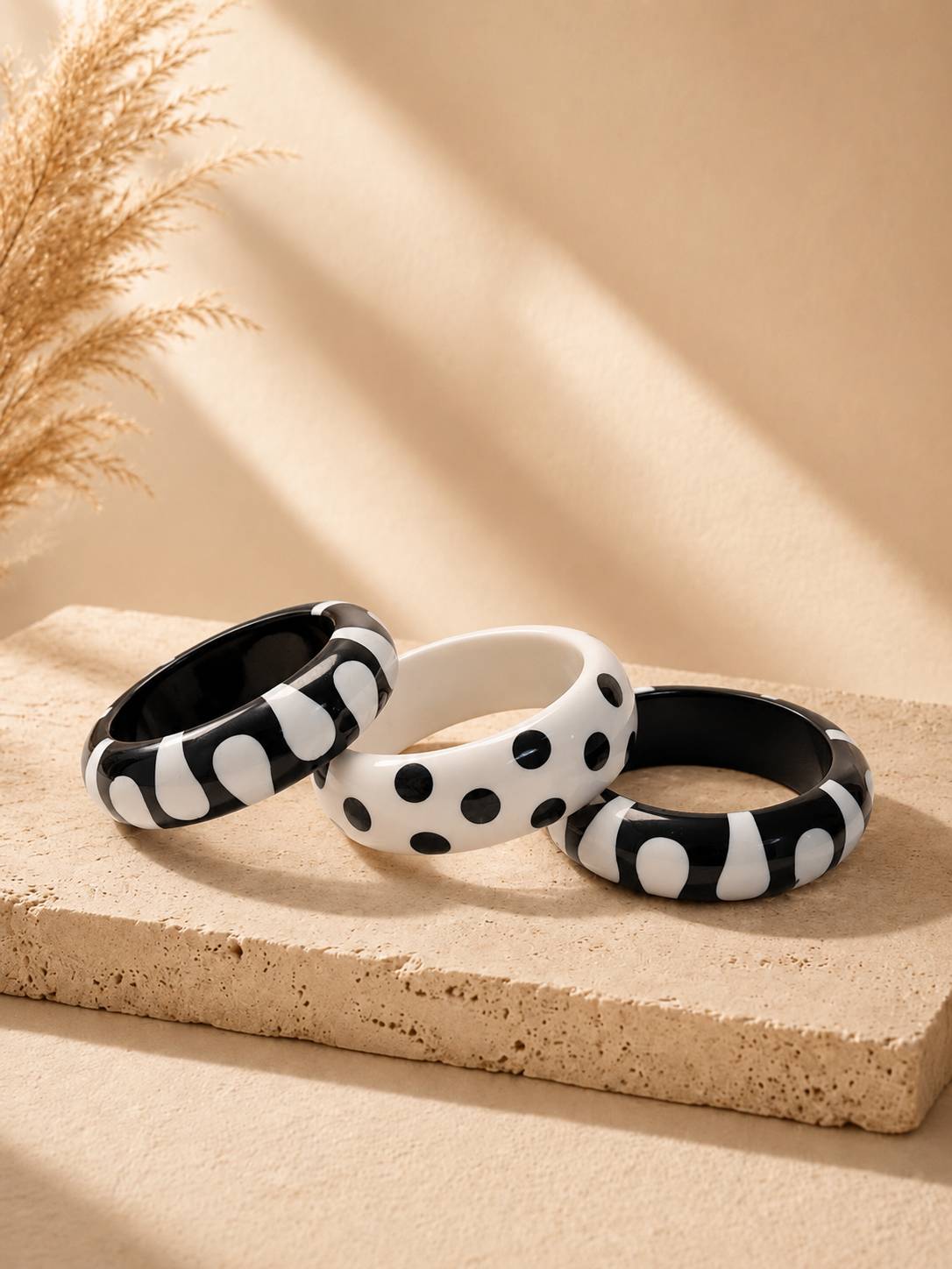 Three black and white resin bangles with blob and polka dot patterns arranged on a beige stone surface with dried pampas grass