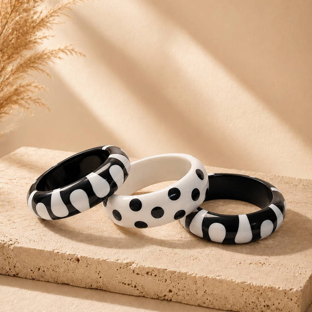 Three black and white resin bangles with blob and polka dot patterns arranged on a beige stone surface with dried pampas grass
