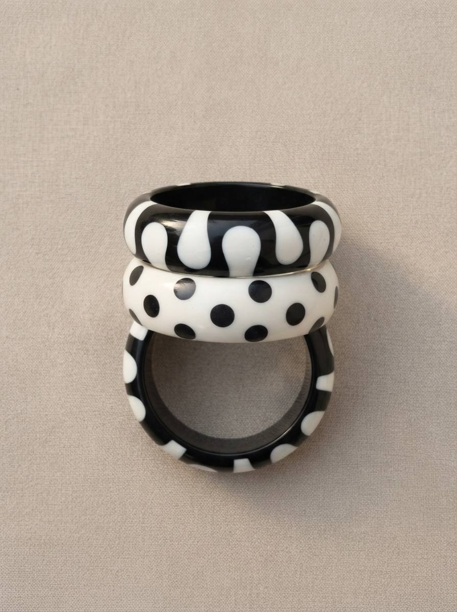 Three black and white resin bangles stacked vertically showing blob, polka dot, and spot patterns against a grey fabric background