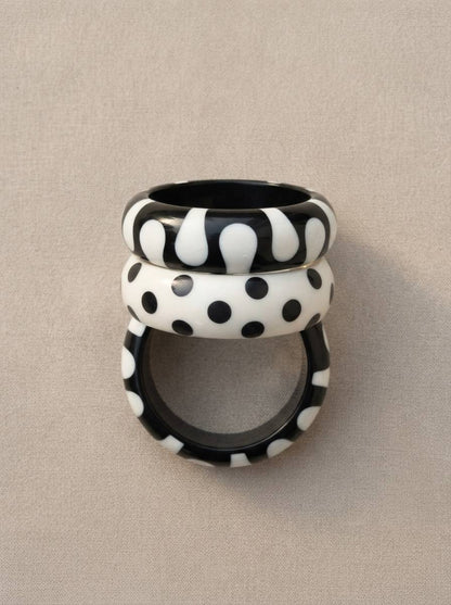 Three black and white resin bangles stacked vertically showing blob, polka dot, and spot patterns against a grey fabric background