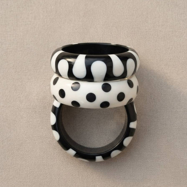 Three black and white resin bangles stacked vertically showing blob, polka dot, and spot patterns against a grey fabric background