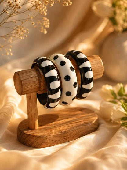 Three black and white resin bangles displayed on a wooden jewellery stand surrounded by dried flowers and white blossoms