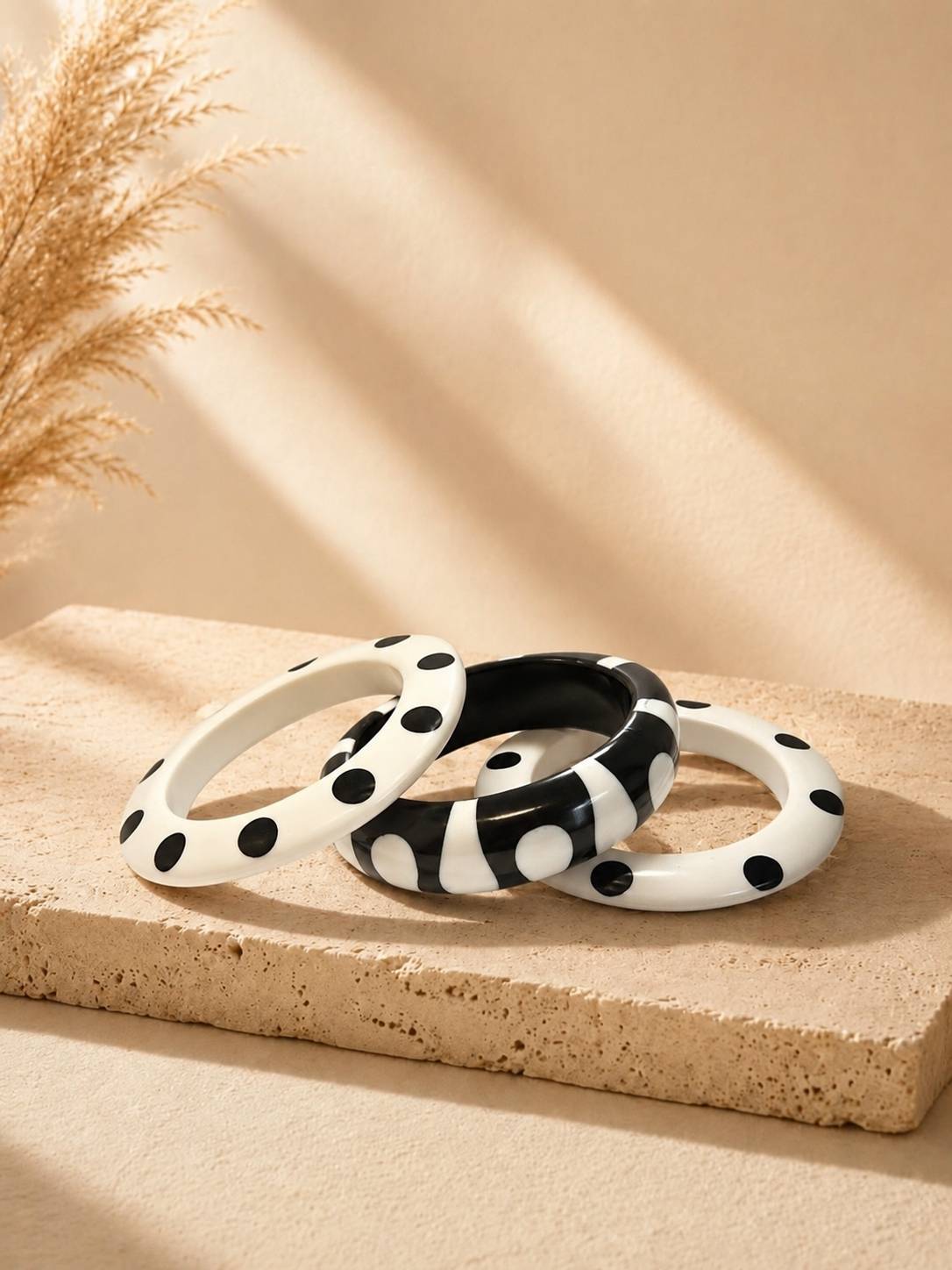 Three black and white polka dot resin bangles stacked on a beige stone surface with dried pampas grass in the background