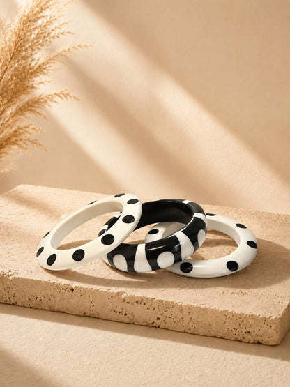 Three black and white polka dot resin bangles stacked on a beige stone surface with dried pampas grass in the background