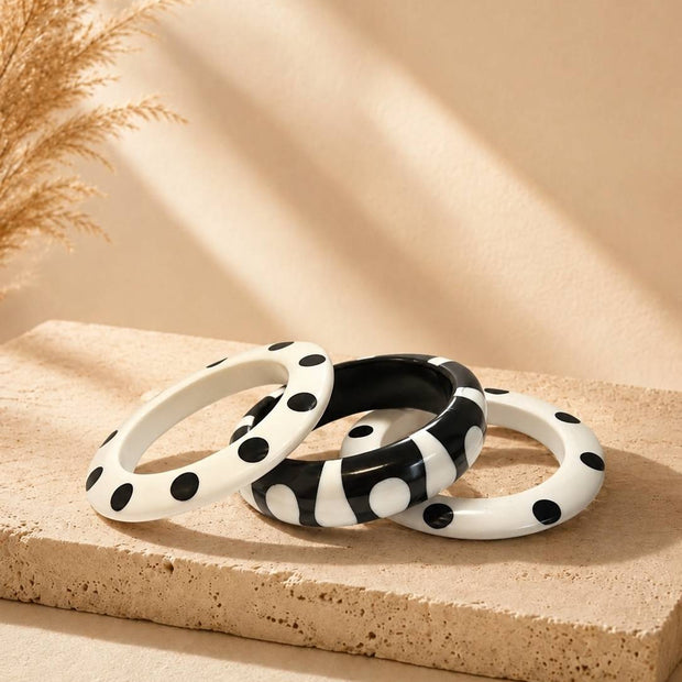 Three black and white polka dot resin bangles stacked on a beige stone surface with dried pampas grass in the background