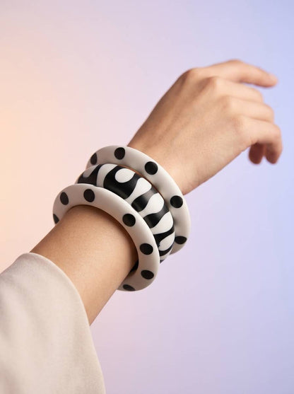 Stack of three black and white resin bangles worn on a raised wrist against a soft lavender and pink gradient background