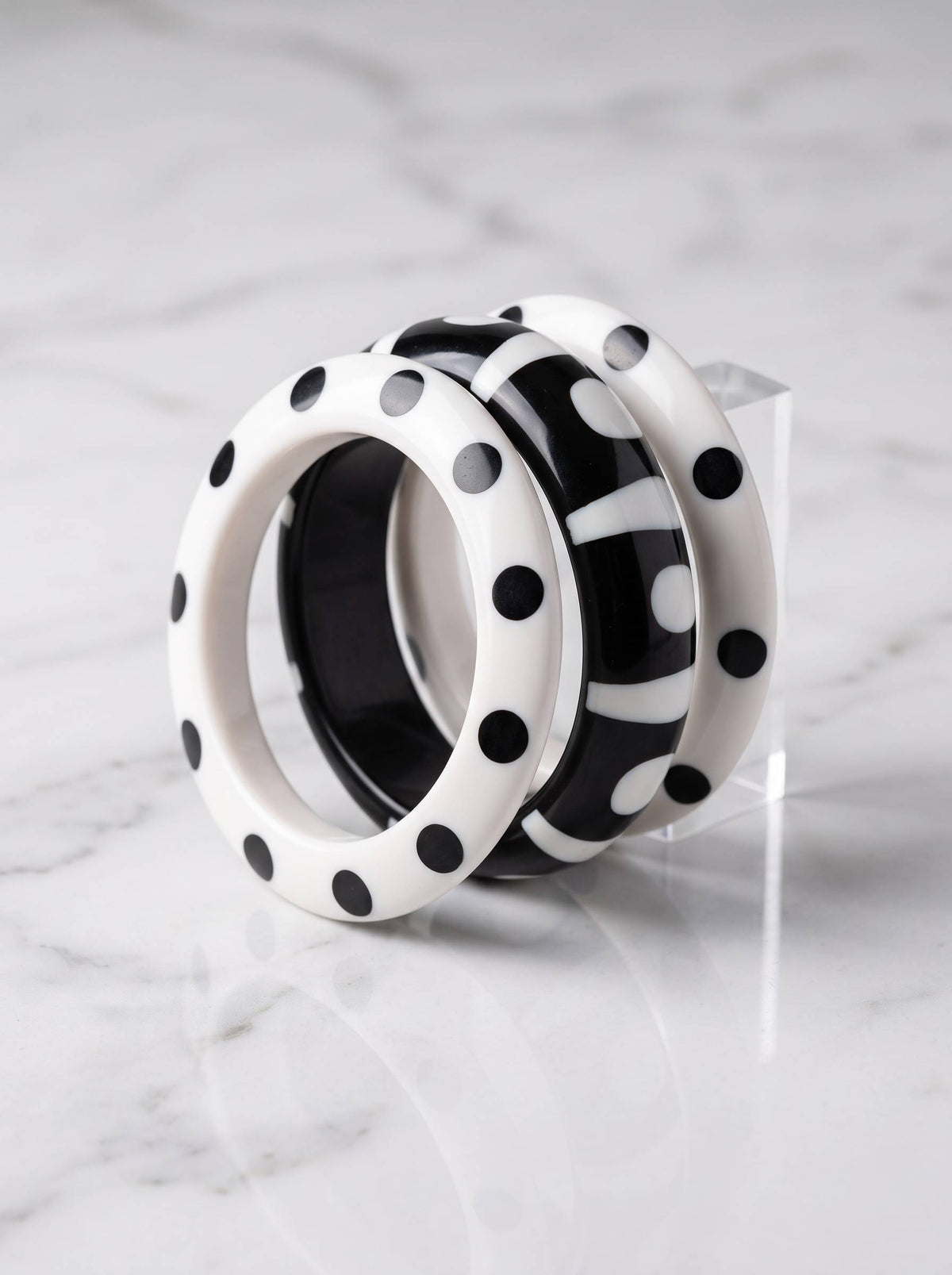Two white polka dot bangles and one black stripe dot bangle propped on a clear acrylic stand against white marble