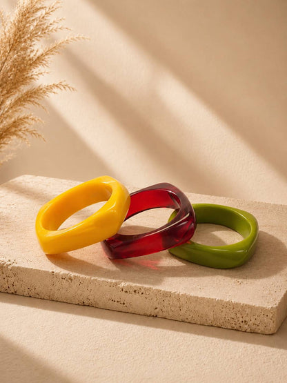 Three colourful resin bangles in yellow, red, and olive green arranged on a beige stone surface with dried pampas grass