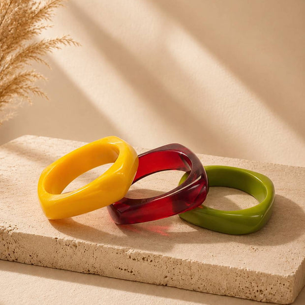 Three colourful resin bangles in yellow, red, and olive green arranged on a beige stone surface with dried pampas grass