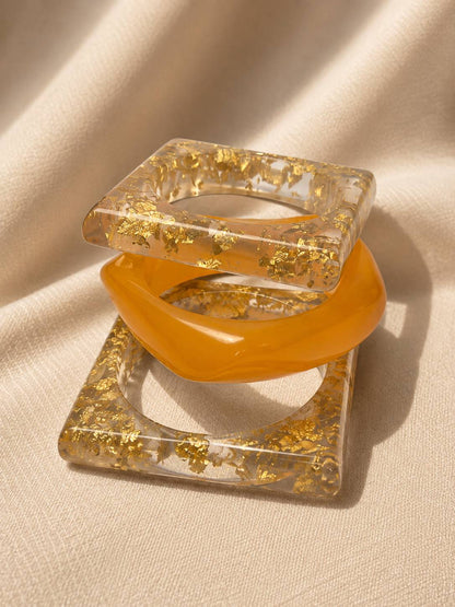 Three resin bangles stacked together on cream fabric, showing square gold foil frames and rounded amber bangle