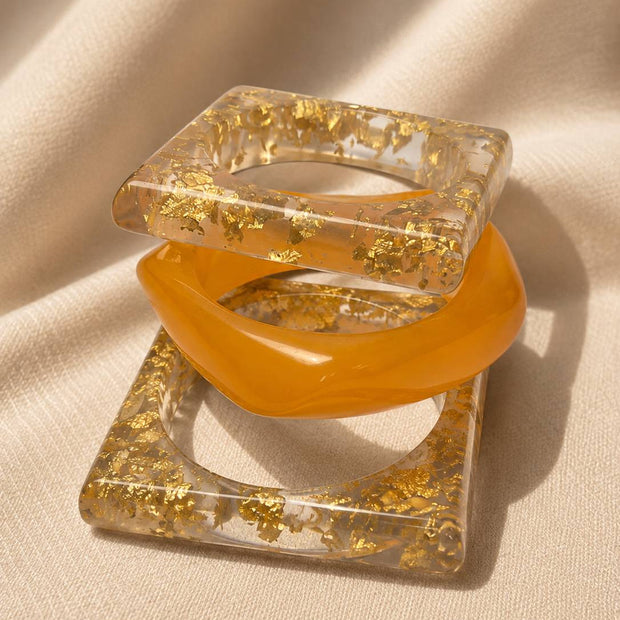 Three resin bangles stacked together on cream fabric, showing square gold foil frames and rounded amber bangle