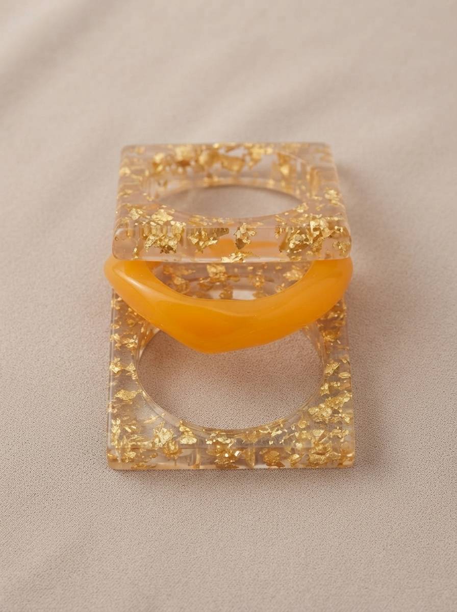 Top view of square gold foil resin bangles and amber dome bangle stacked flat on beige surface